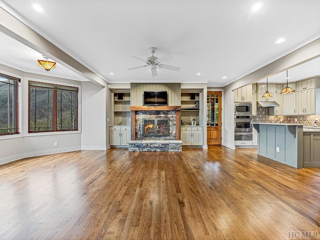 unfurnished living room featuring crown molding, ceiling fan, a fireplace, built in shelves, and light wood-type flooring