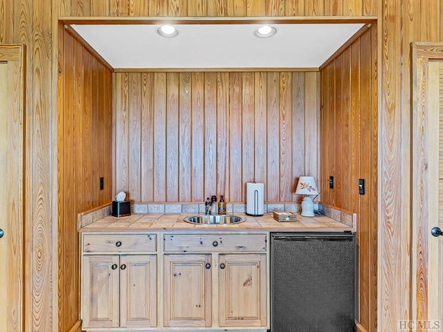 bar featuring light brown cabinetry, sink, tile countertops, wooden walls, and dishwasher