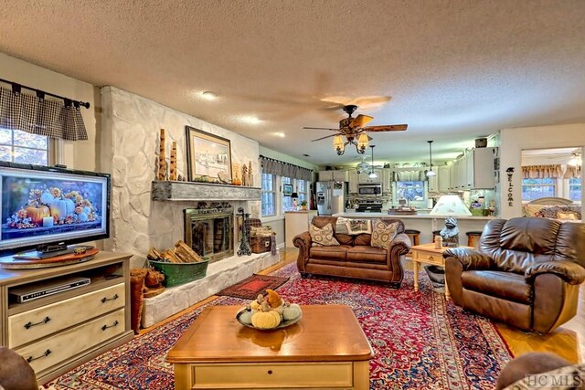 living room featuring ceiling fan, a stone fireplace, wood-type flooring, and a textured ceiling