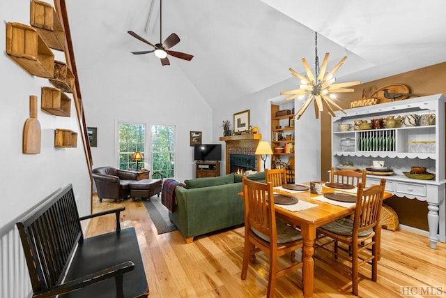 dining space with ceiling fan with notable chandelier, a fireplace, high vaulted ceiling, and light hardwood / wood-style flooring