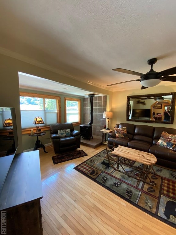 living room with crown molding, a textured ceiling, a wood stove, ceiling fan, and light hardwood / wood-style floors