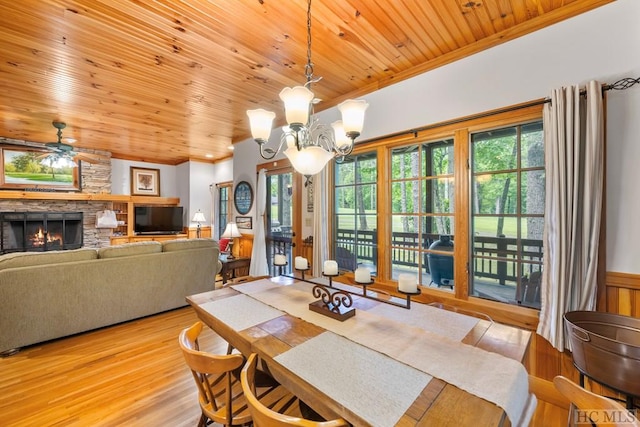 dining space with a stone fireplace, light hardwood / wood-style flooring, wooden ceiling, ornamental molding, and ceiling fan with notable chandelier