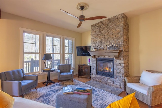 living area featuring baseboards, a stone fireplace, wood finished floors, and a ceiling fan