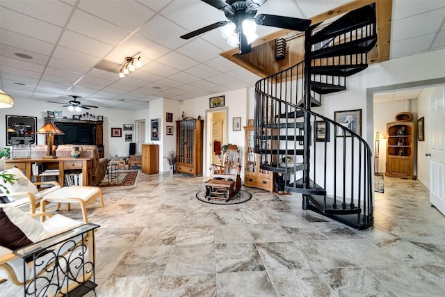 living room featuring a ceiling fan, a drop ceiling, and stairs