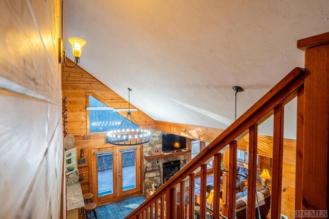 staircase with wooden walls, a stone fireplace, a chandelier, and lofted ceiling