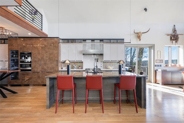 kitchen with light wood-style flooring, a towering ceiling, white cabinetry, modern cabinets, and tasteful backsplash