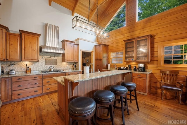 kitchen featuring wall chimney exhaust hood, a breakfast bar, stainless steel gas cooktop, a center island with sink, and pendant lighting