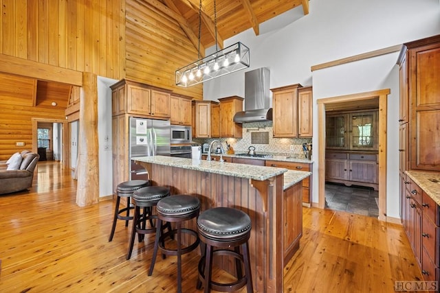 kitchen featuring light stone counters, appliances with stainless steel finishes, wall chimney exhaust hood, and an island with sink