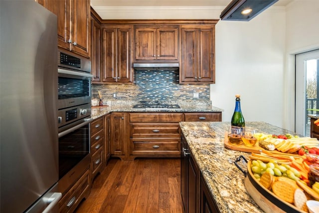 kitchen featuring appliances with stainless steel finishes, dark wood finished floors, backsplash, under cabinet range hood, and light stone counters