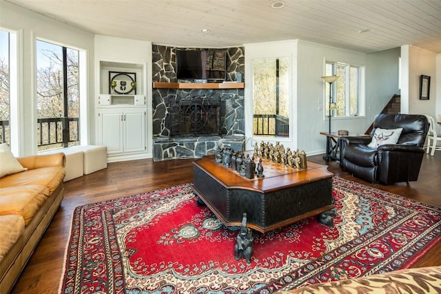 living area with wood finished floors, wooden ceiling, healthy amount of natural light, ornamental molding, and a stone fireplace