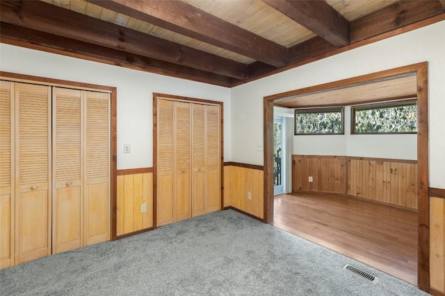 unfurnished bedroom with wood walls, wooden ceiling, carpet floors, two closets, and beam ceiling