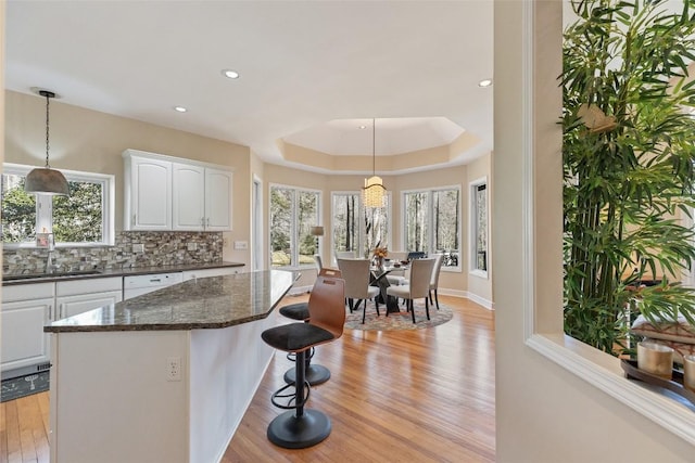 kitchen featuring light wood-style flooring, white cabinets, backsplash, dark stone counters, and a tray ceiling