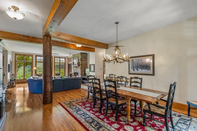 dining room featuring beamed ceiling, wood-type flooring, a stone fireplace, and a notable chandelier