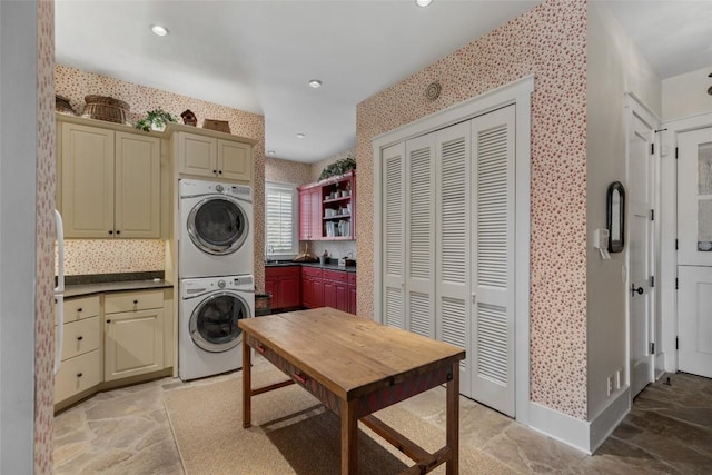 clothes washing area featuring stacked washer and clothes dryer, stone finish floor, wallpapered walls, recessed lighting, and cabinet space