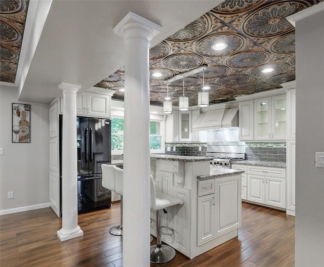 kitchen featuring custom range hood, white cabinetry, black fridge, decorative backsplash, and decorative columns