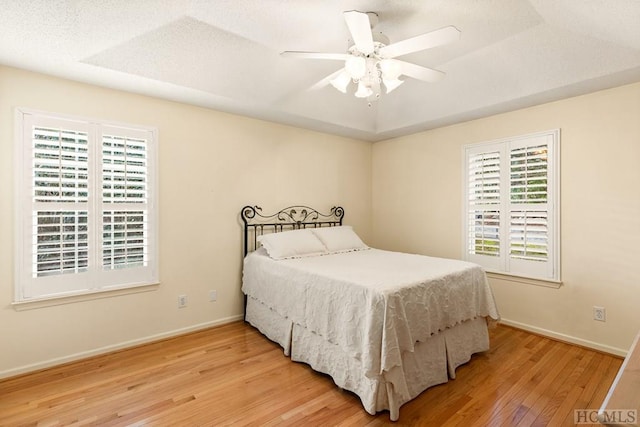 bedroom with multiple windows, wood-type flooring, ceiling fan, and a tray ceiling