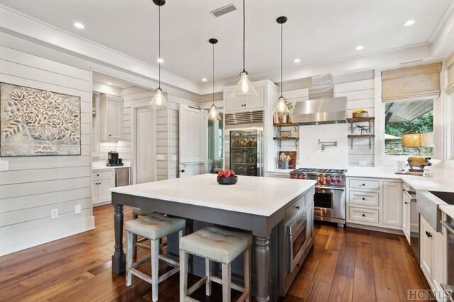 kitchen featuring wall chimney range hood, built in appliances, white cabinets, and a kitchen breakfast bar