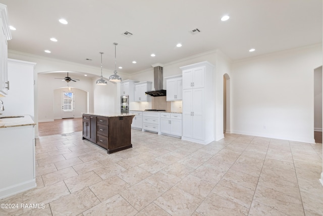 kitchen with wall chimney exhaust hood, ceiling fan, pendant lighting, white cabinetry, and an island with sink