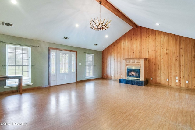 unfurnished living room featuring light hardwood / wood-style floors, beamed ceiling, wooden walls, a tiled fireplace, and an inviting chandelier