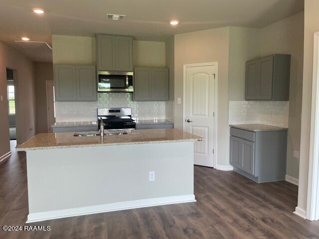 kitchen with dark wood-type flooring, gray cabinetry, appliances with stainless steel finishes, light stone countertops, and a kitchen island with sink