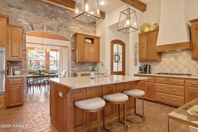 kitchen featuring custom range hood, backsplash, an island with sink, beam ceiling, and gas stovetop