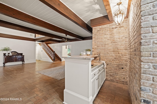 kitchen with hanging light fixtures, white cabinets, beamed ceiling, ceiling fan with notable chandelier, and brick wall