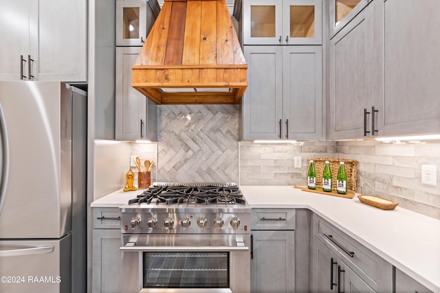 kitchen with gray cabinetry, custom range hood, backsplash, and appliances with stainless steel finishes