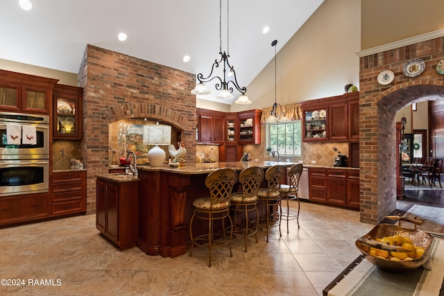 kitchen featuring pendant lighting, a center island with sink, tasteful backsplash, light stone counters, and stainless steel double oven