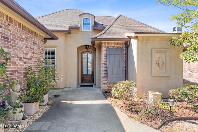 doorway to property featuring roof with shingles, brick siding, and stucco siding