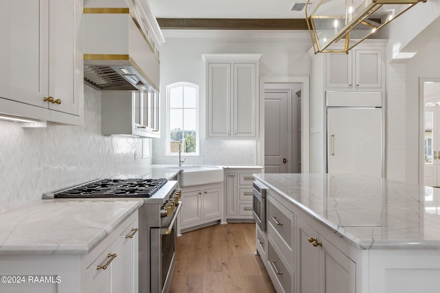 kitchen with light stone countertops, custom exhaust hood, sink, built in appliances, and hanging light fixtures