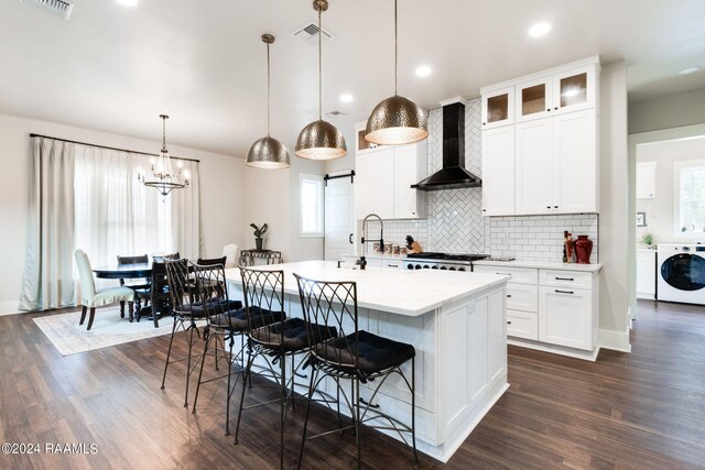 kitchen featuring decorative light fixtures, washer / clothes dryer, an island with sink, white cabinetry, and wall chimney exhaust hood