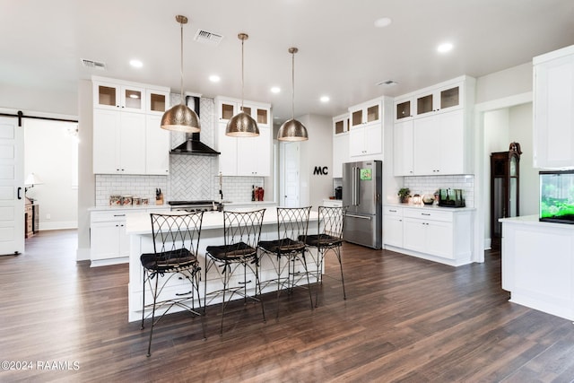 kitchen with high end refrigerator, decorative light fixtures, a center island with sink, a barn door, and wall chimney range hood