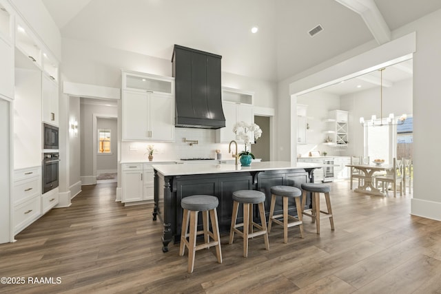 kitchen with an island with sink, custom range hood, black appliances, high vaulted ceiling, and white cabinetry