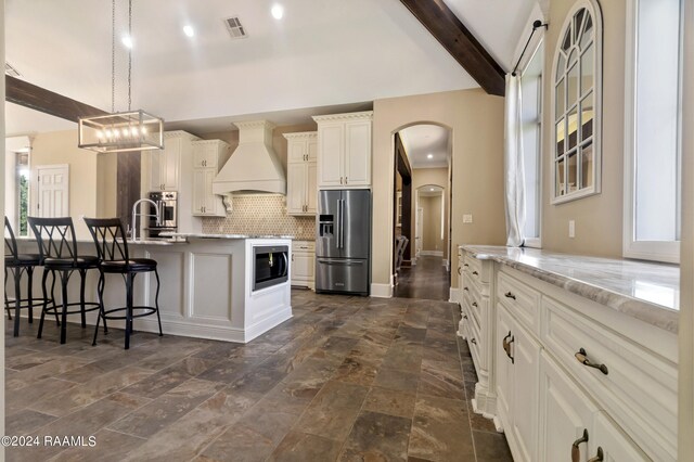 kitchen featuring custom range hood, appliances with stainless steel finishes, light stone counters, beamed ceiling, and a center island with sink