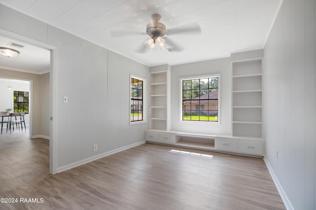 empty room featuring ceiling fan, built in shelves, wood finished floors, baseboards, and crown molding