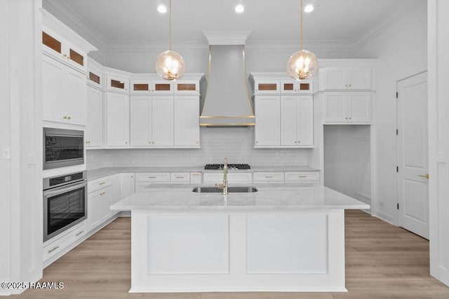 kitchen with built in microwave, an island with sink, a sink, premium range hood, and stainless steel oven