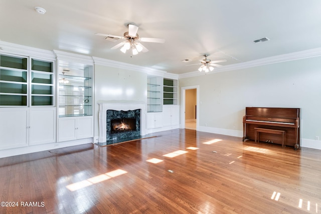 unfurnished living room with built in shelves, ceiling fan, ornamental molding, a high end fireplace, and wood-type flooring