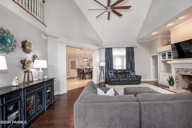 living room featuring high vaulted ceiling, dark wood-type flooring, a fireplace, a ceiling fan, and decorative columns