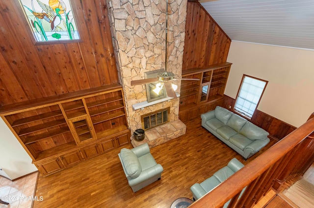 living room featuring wood walls, light hardwood / wood-style flooring, high vaulted ceiling, a stone fireplace, and ceiling fan