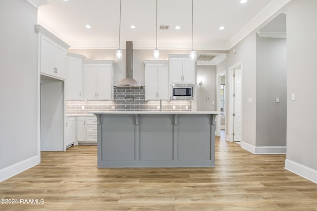 kitchen with light hardwood / wood-style floors, stainless steel microwave, wall chimney exhaust hood, and an island with sink