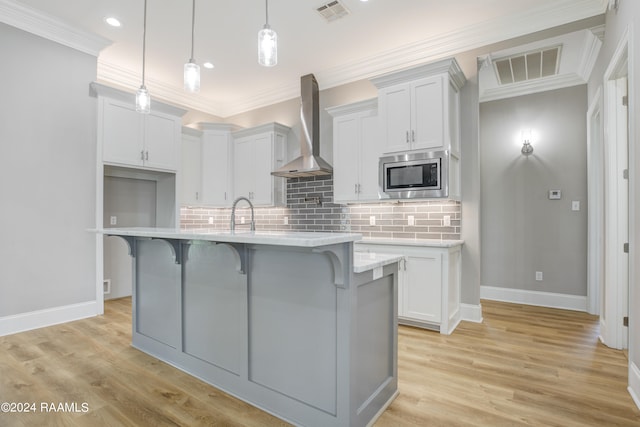 kitchen with pendant lighting, an island with sink, wall chimney exhaust hood, white cabinetry, and stainless steel microwave