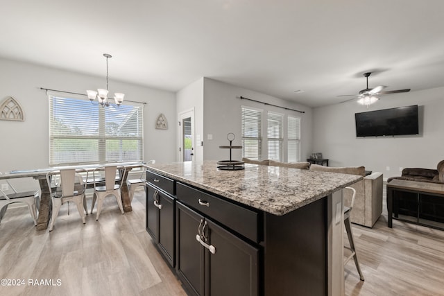 kitchen with light stone counters, ceiling fan with notable chandelier, a kitchen island, pendant lighting, and light hardwood / wood-style flooring