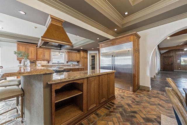 kitchen with built in fridge, a tray ceiling, a kitchen bar, premium range hood, and ornamental molding