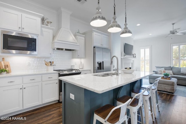 kitchen with an island with sink, white cabinetry, custom exhaust hood, stainless steel appliances, and decorative light fixtures