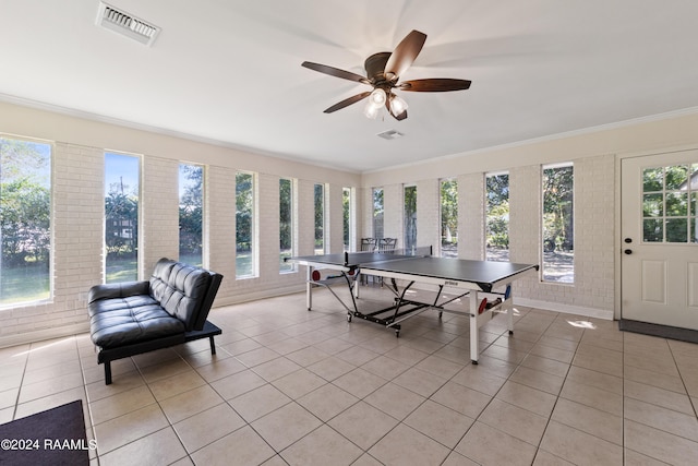 rec room with light tile patterned floors, ceiling fan, ornamental molding, and brick wall