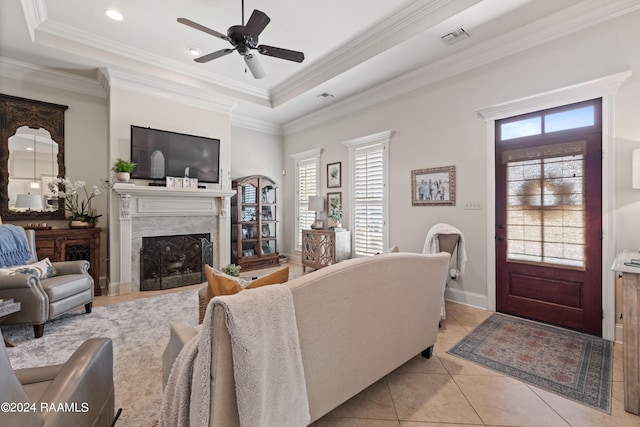 tiled living room with ornamental molding, a premium fireplace, a tray ceiling, and ceiling fan