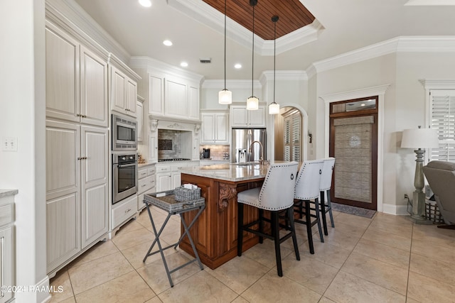 kitchen featuring an island with sink, stainless steel appliances, pendant lighting, light stone counters, and ornamental molding