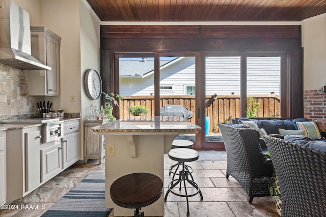 kitchen with wall chimney range hood, wood ceiling, a center island, and a kitchen bar