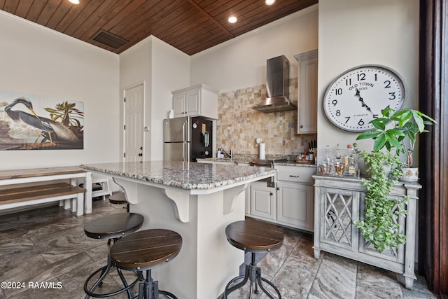 kitchen with wall chimney range hood, a breakfast bar, wood ceiling, and stainless steel refrigerator