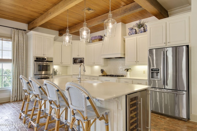 kitchen with beam ceiling, a center island with sink, decorative light fixtures, wine cooler, and stainless steel appliances